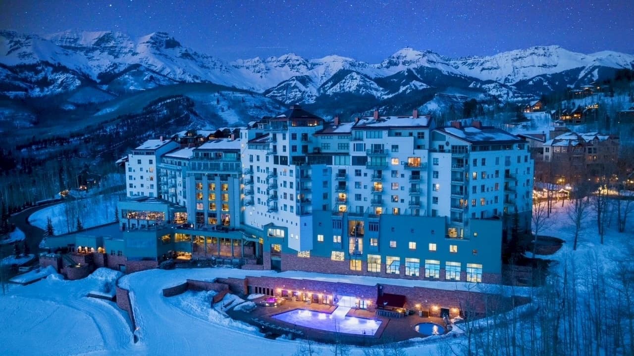 The Peaks Resort from above on a winter night, its windows lit and the heated outdoor pool glowing, with snowy peaks behind.