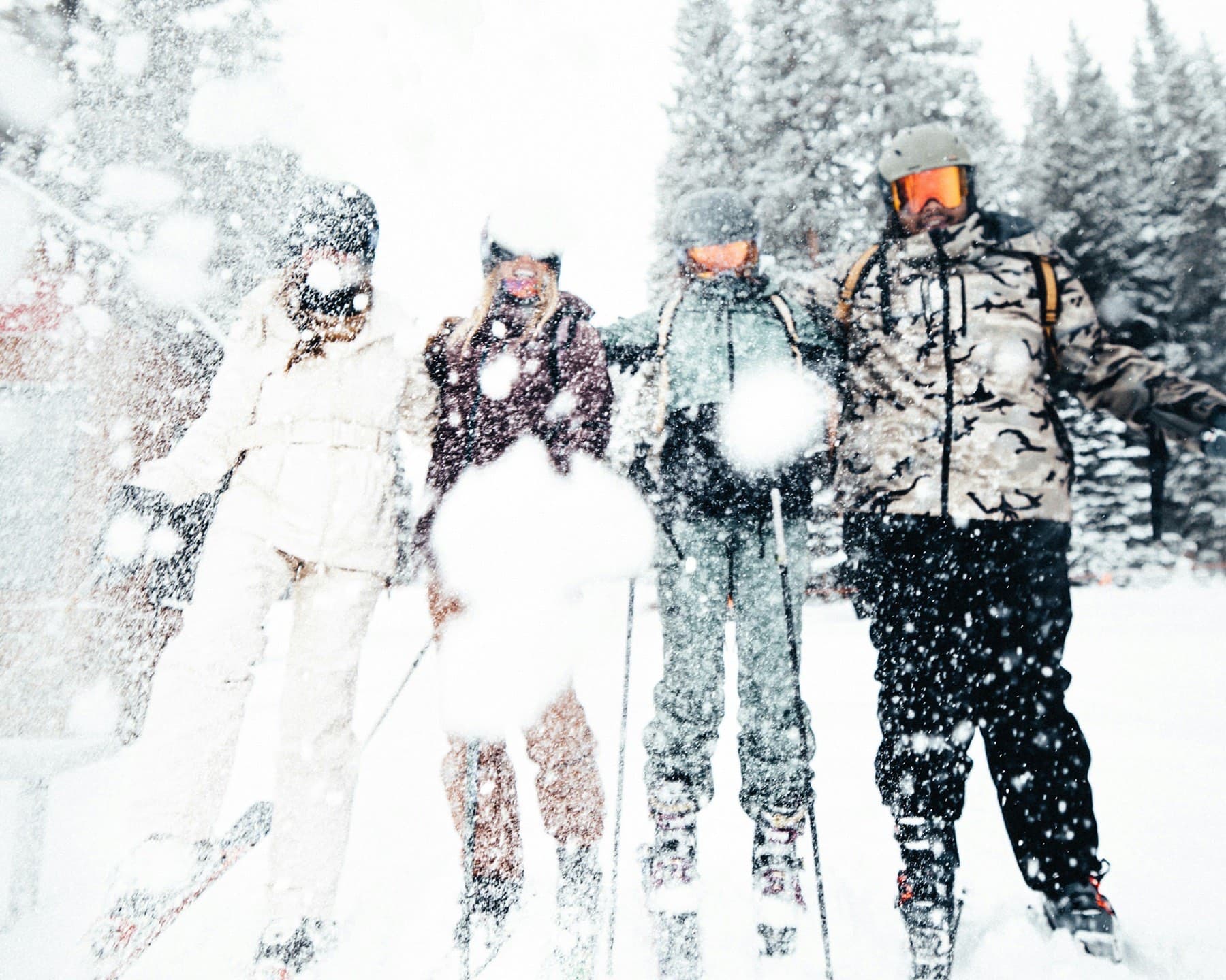 Four friends on skis, arms linked, laughing as someone throws a handful of powder at them in falling snow.